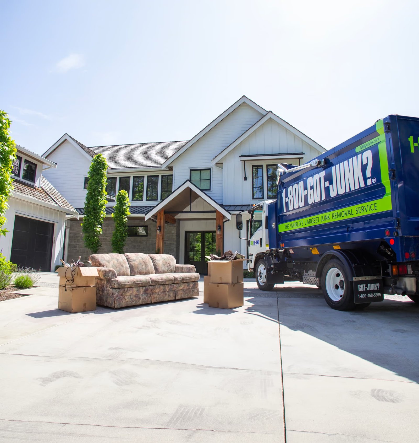 Blue 1-800-GOT-JUNK? truck parked in front of a large white house, with a couch and cardboard boxes on the driveway