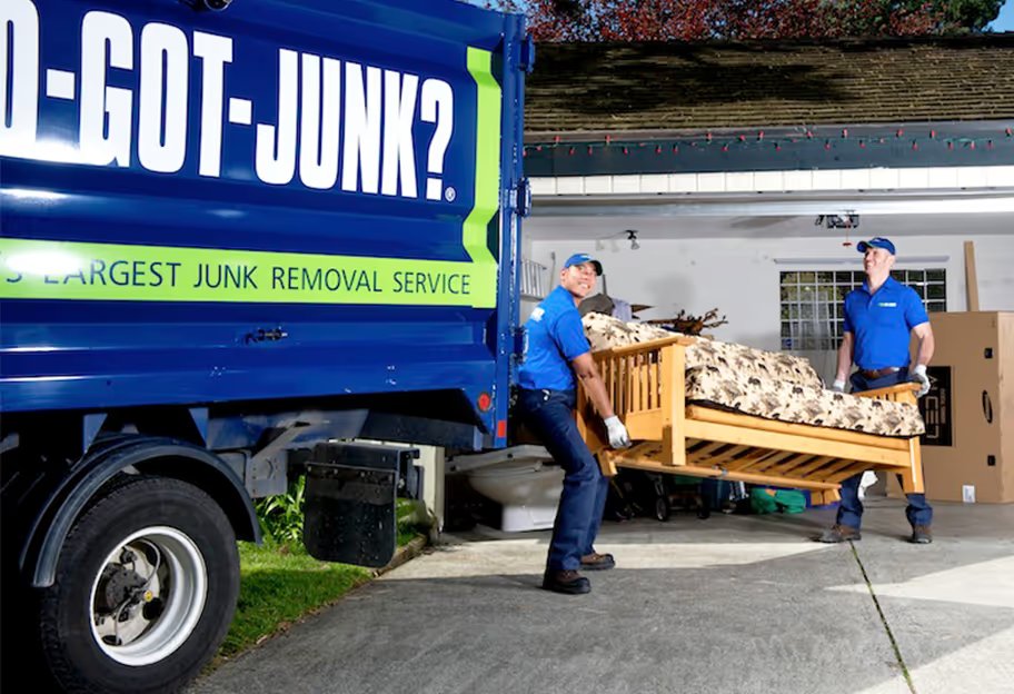 Two uniformed crew members carry a wooden futon frame toward a blue 1-800-GOT-JUNK? truck parked in a driveway
