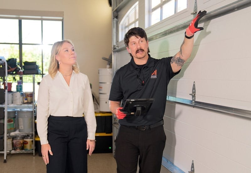 An A1 technician holding a tablet points to the track of a garage door while a woman stands beside him in the garage