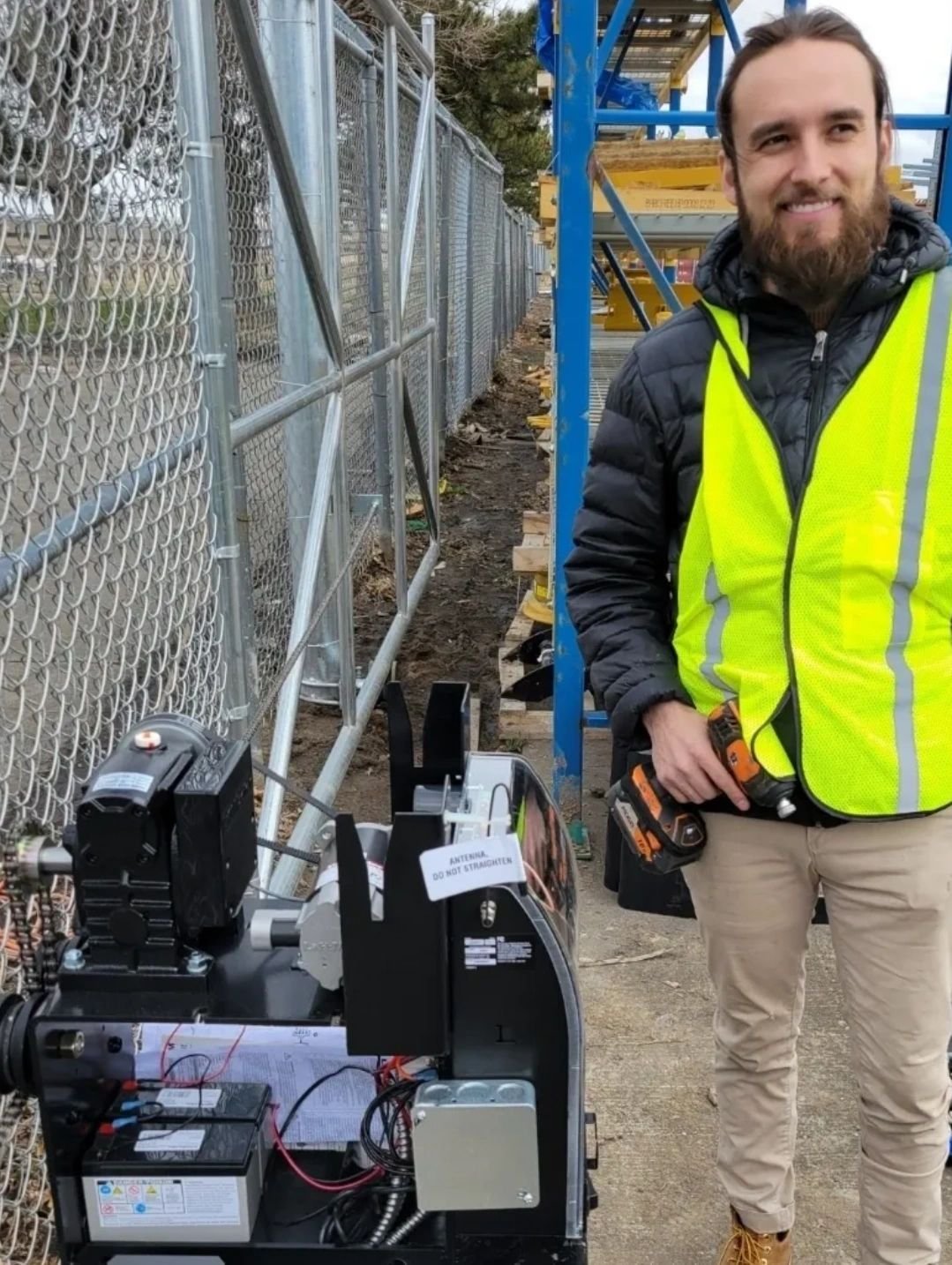 Smiling bearded man in a yellow safety vest stands beside an automatic gate operator with batteries and wiring next to chain-link fence