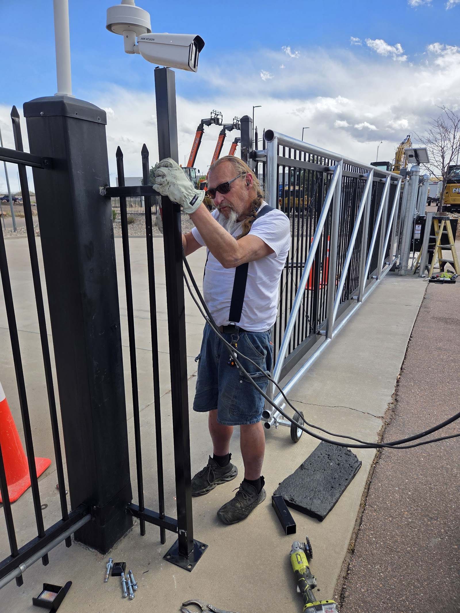 Worker in gloves and sunglasses runs cable along a black iron fence post beside a large rolling gate and security camera