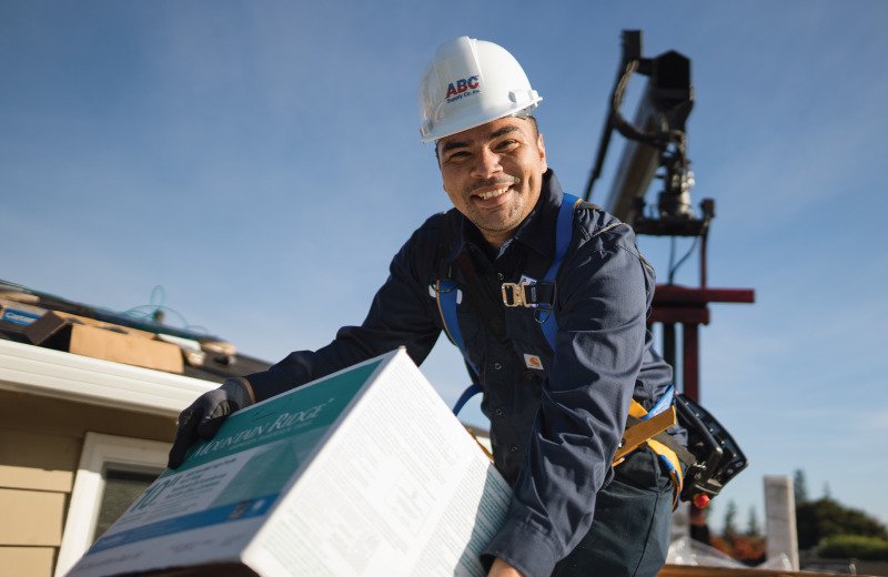 Smiling worker in an ABC hard hat and safety harness lifts a box of roofing material on a roof