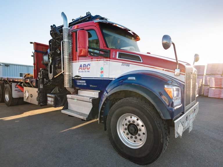 Front view of a red, white and blue ABC Supply delivery truck with a boom crane parked beside stacked building materials