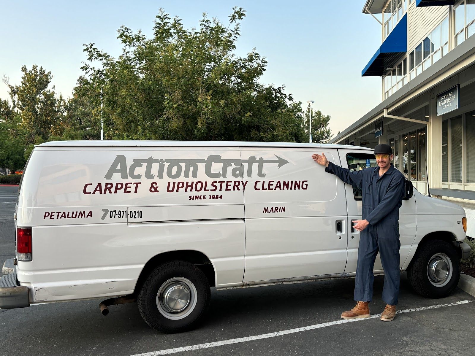 A white cargo van lettered Action Craft Carpet & Upholstery Cleaning, Since 1984, with Petaluma and Marin on its side, as a man in coveralls gestures toward it