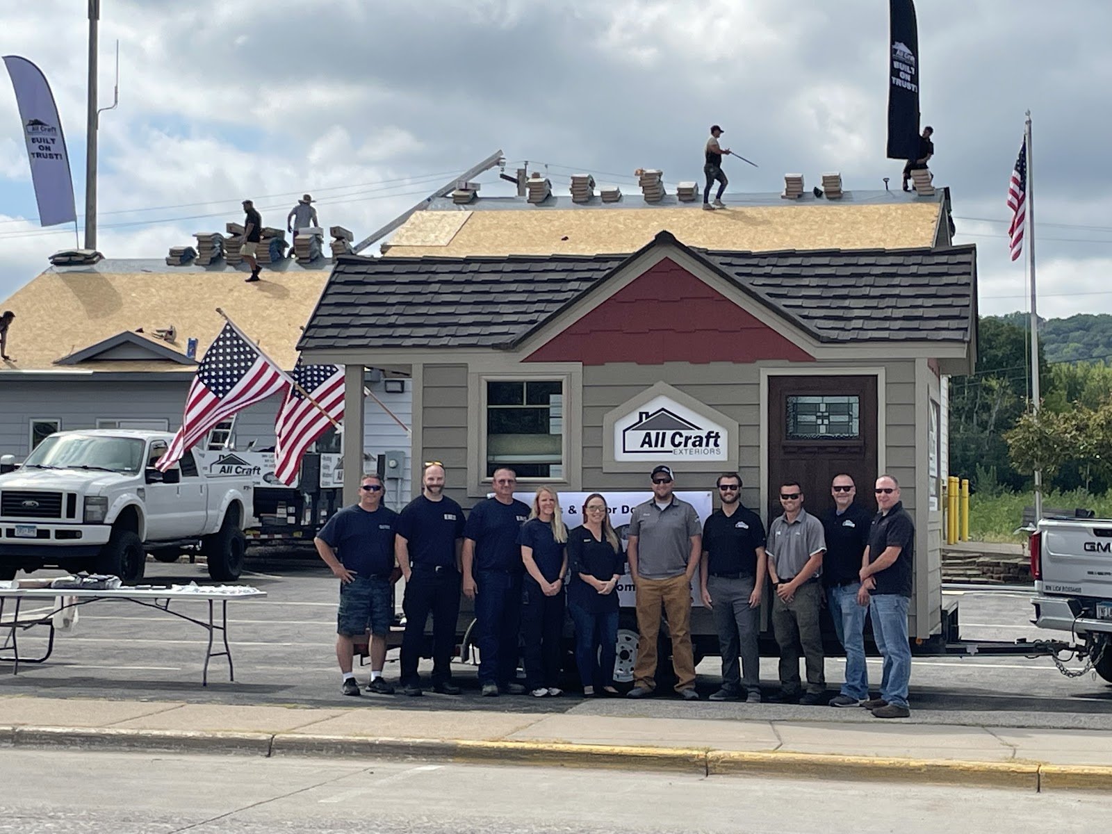 Ten All Craft Exteriors staff members stand in front of a trailer-mounted display building bearing the company logo while roofers work on a roof behind them