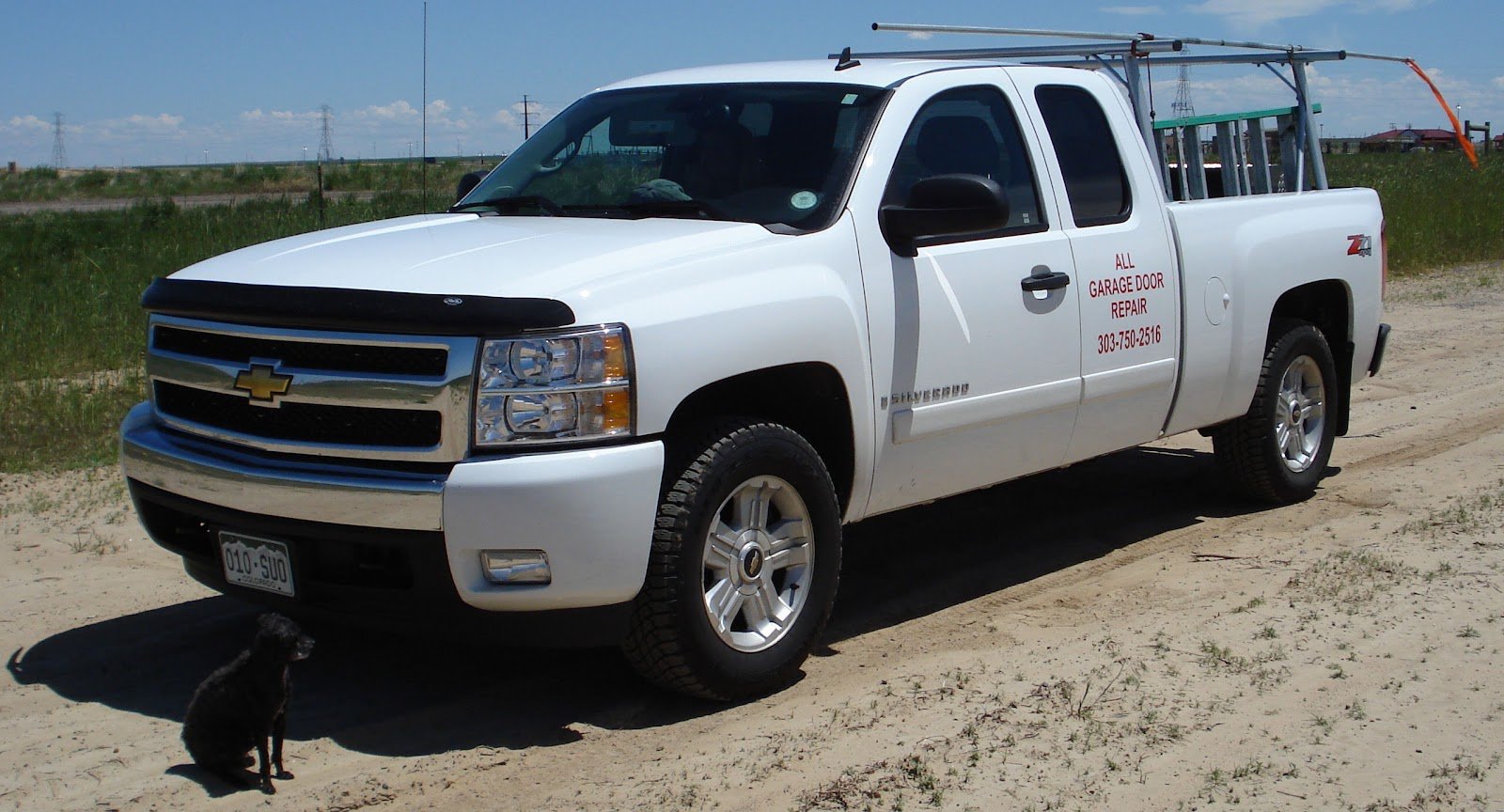 A white Chevrolet pickup with a ladder rack, lettered for All Garage Door Repair, parked on a dirt lot with a small black dog sitting by the front bumper