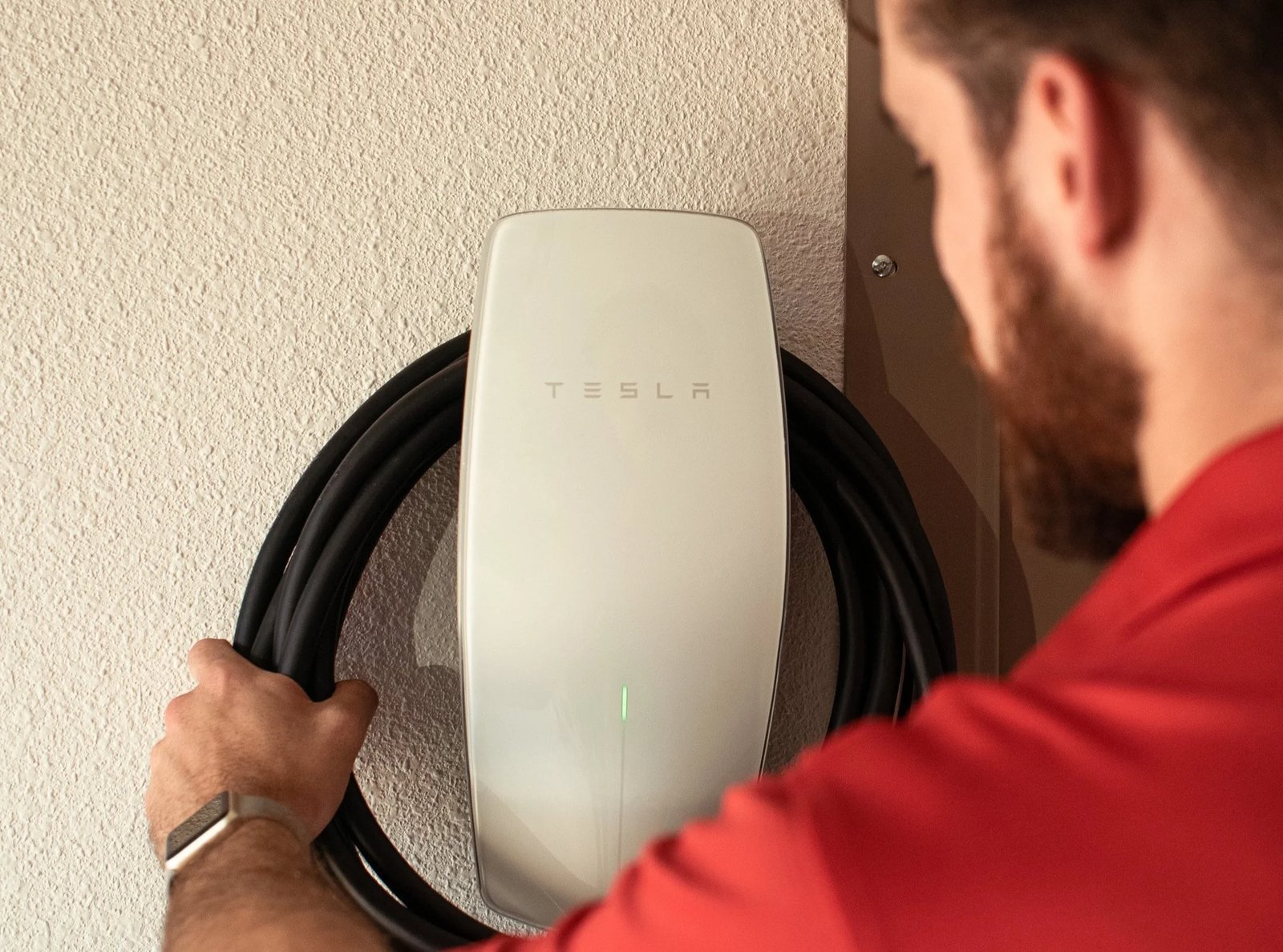 Electrician in a red shirt handles the coiled cable of a white Tesla Wall Connector mounted on a textured wall.