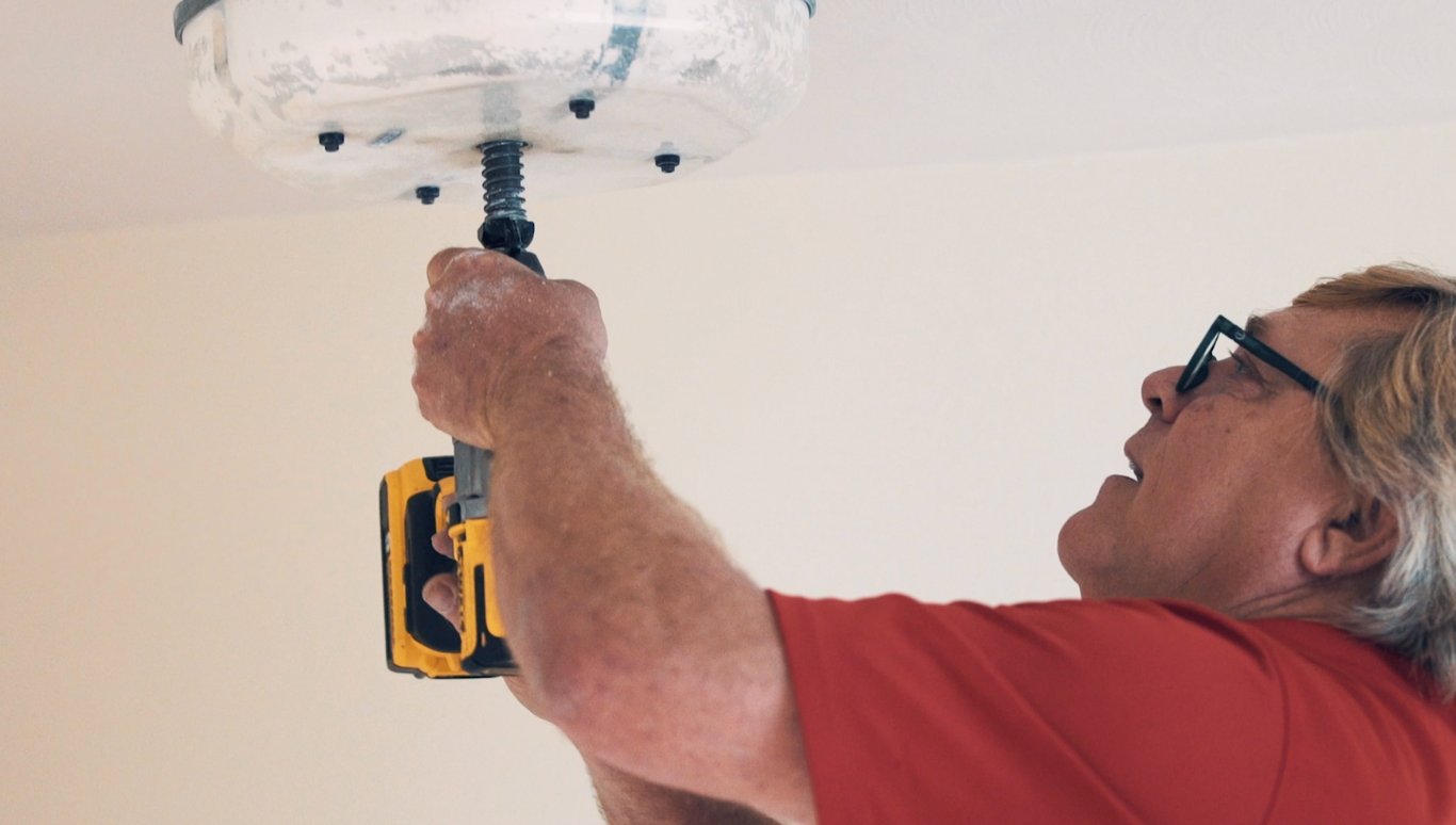 Man with glasses in a red shirt uses a yellow cordless drill to fasten a light fixture to a ceiling.