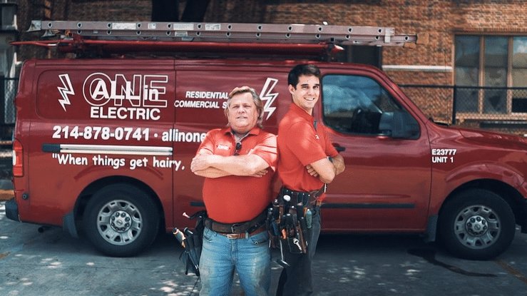 Two men in red All-In-One Electric shirts and tool belts stand back to back, arms crossed, in front of a red company van.