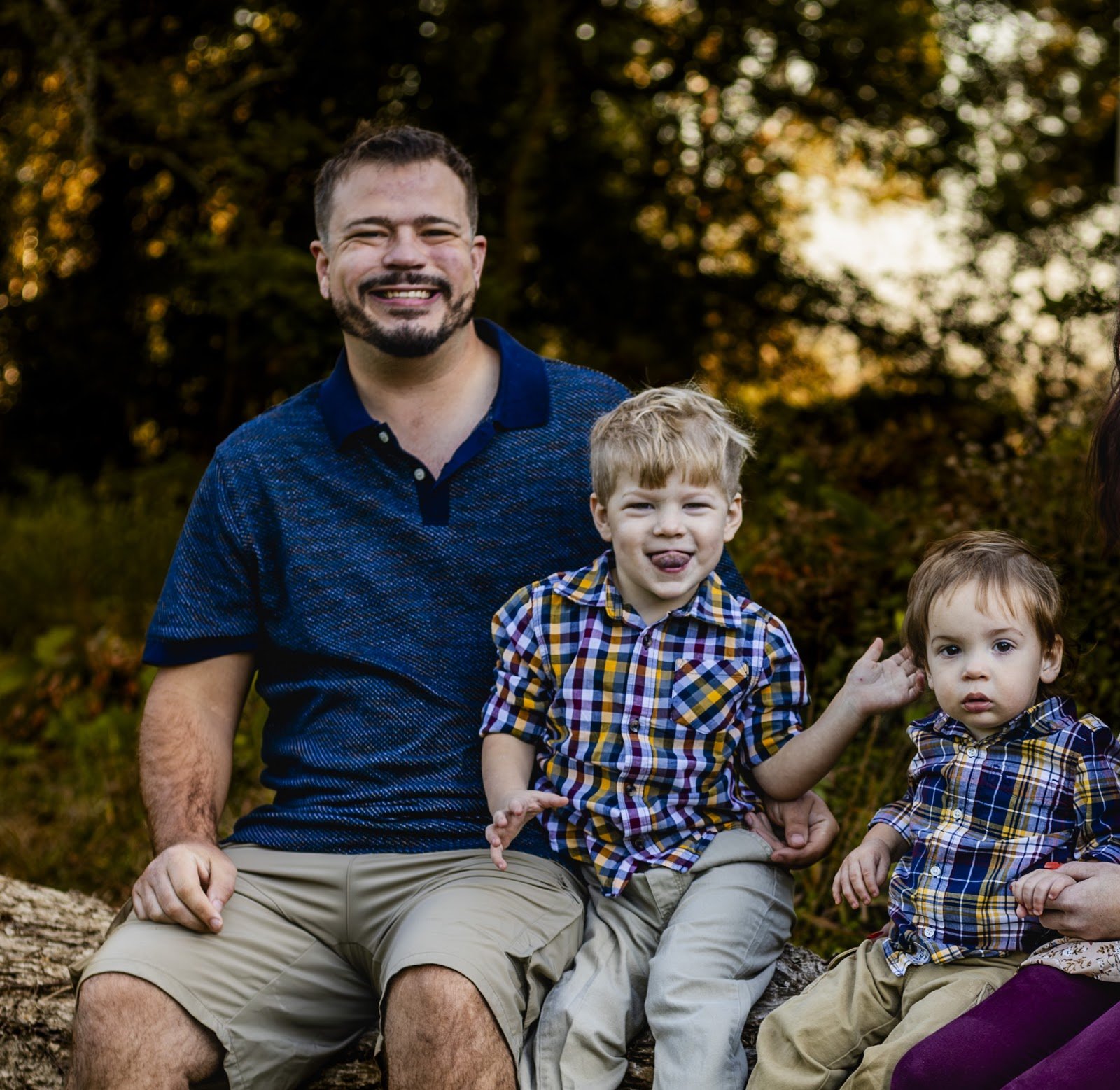 Smiling bearded man in a blue polo sitting outdoors with two young boys in plaid shirts, one sticking out his tongue