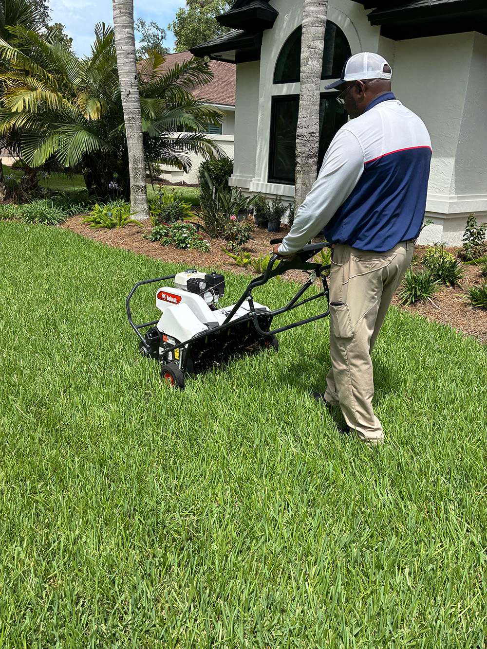 Technician in a navy and white uniform and cap pushes a lawn aerator across a green lawn beside a stucco house