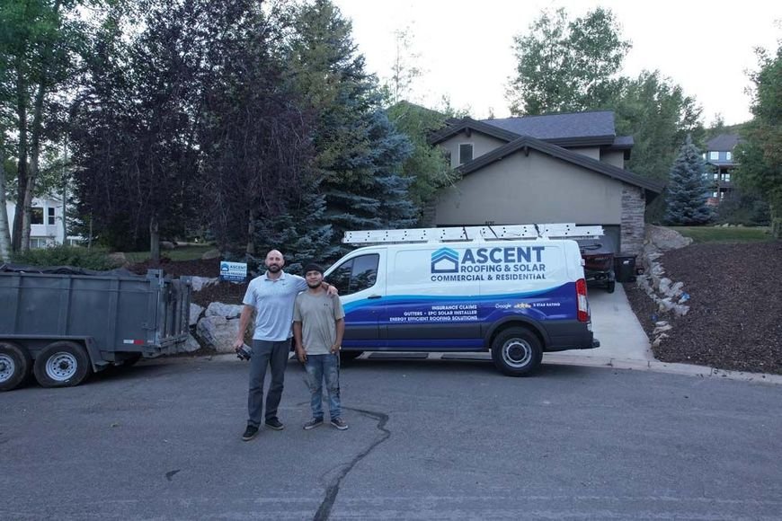 Two men stand in front of a white Ascent Roofing & Solar van with a ladder rack, beside a dump trailer.