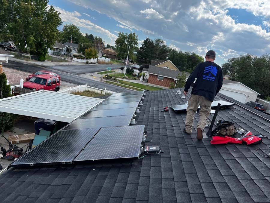 Worker in a dark Ascent shirt stands on a shingled roof beside rows of installed solar panels and tools.