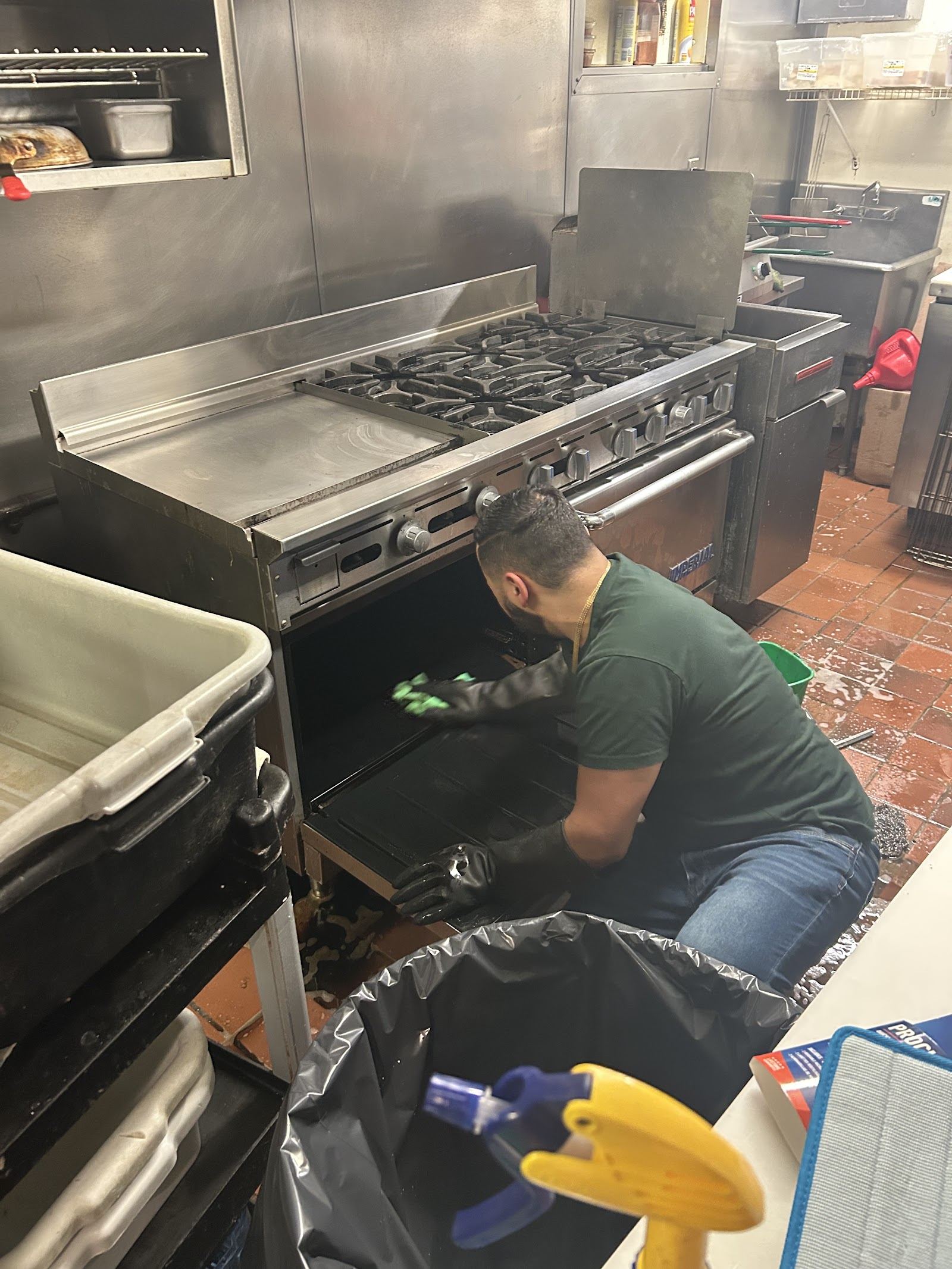 A worker in gloves kneels and scrubs inside the open oven of a commercial range in a restaurant kitchen