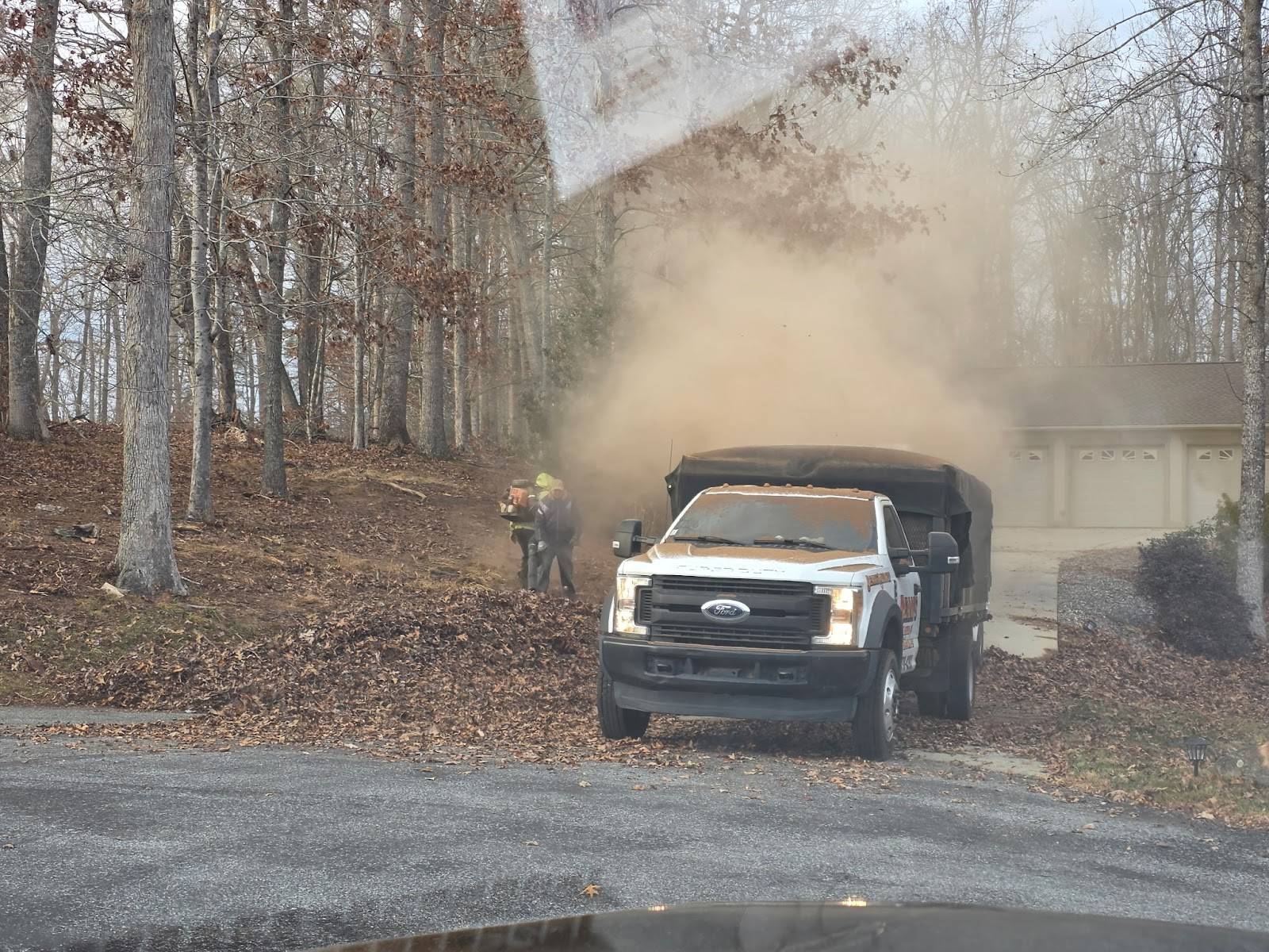 A white work truck with a tarp-covered bed in a wooded driveway, a cloud of leaf dust rising as two workers with backpack blowers clear leaves
