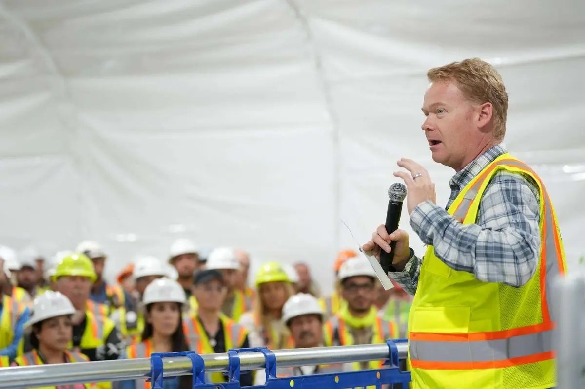 Man in a plaid shirt and safety vest speaks into a microphone to a crowd of hard-hatted workers