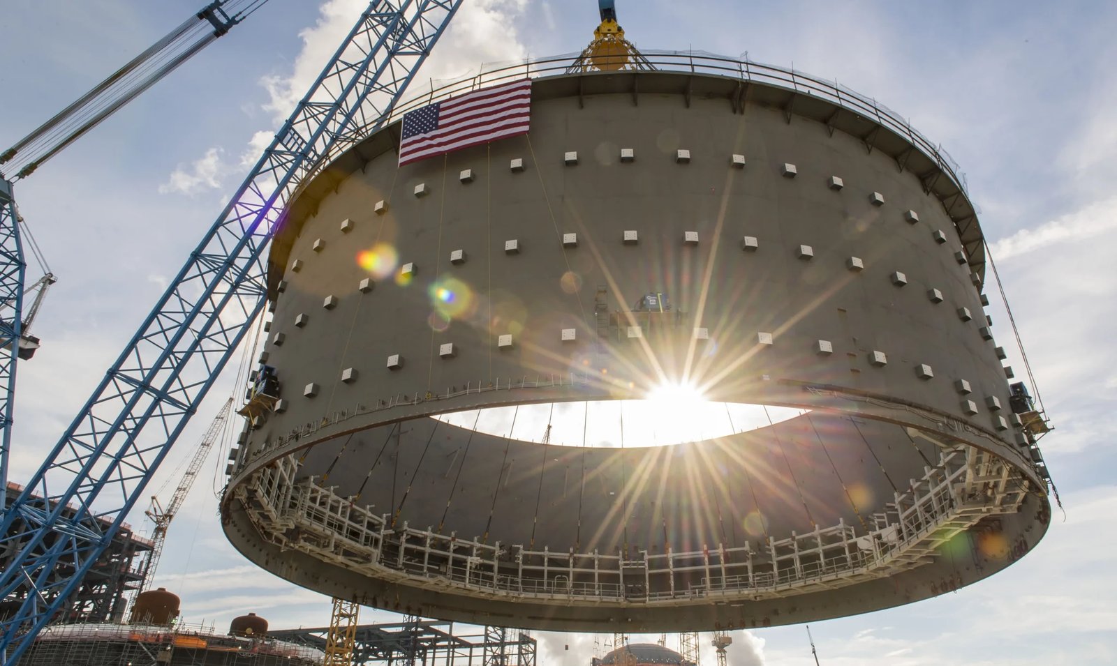 Crane lifts a huge cylindrical steel containment section with an American flag on top, sun shining through the opening