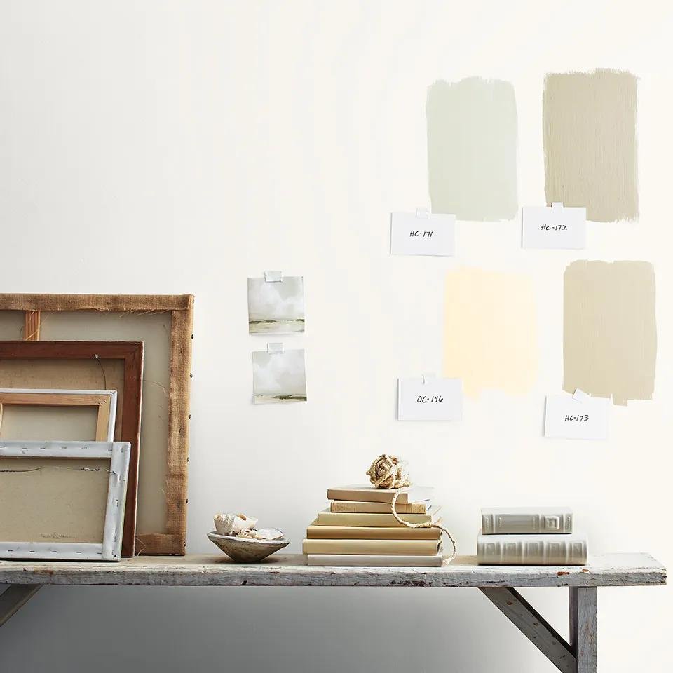 Four painted color swatches with handwritten code labels taped to a white wall above a bench with books and frames
