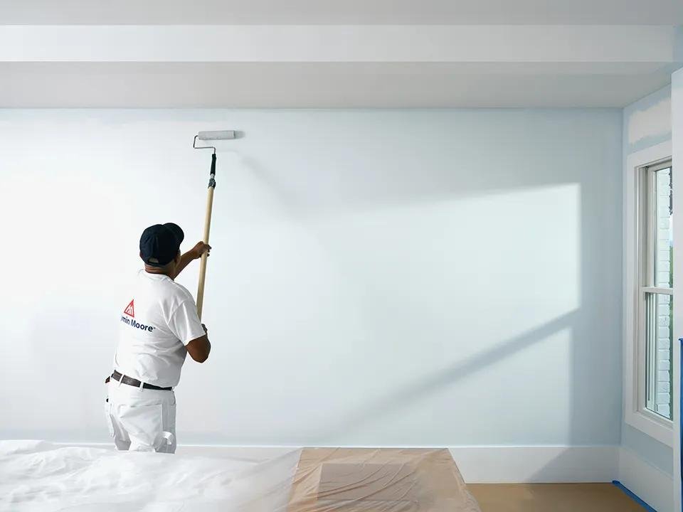 Painter in a white Benjamin Moore shirt and cap rolls pale blue paint onto a wall in a room with drop cloths on the floor