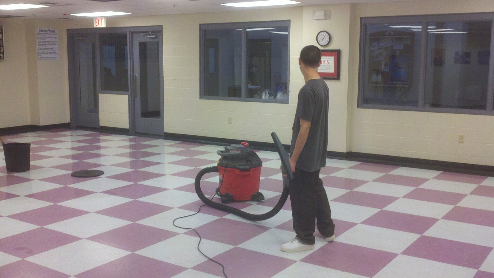 Worker stands with a red wet/dry shop vacuum on a large checkered tile floor in a hallway