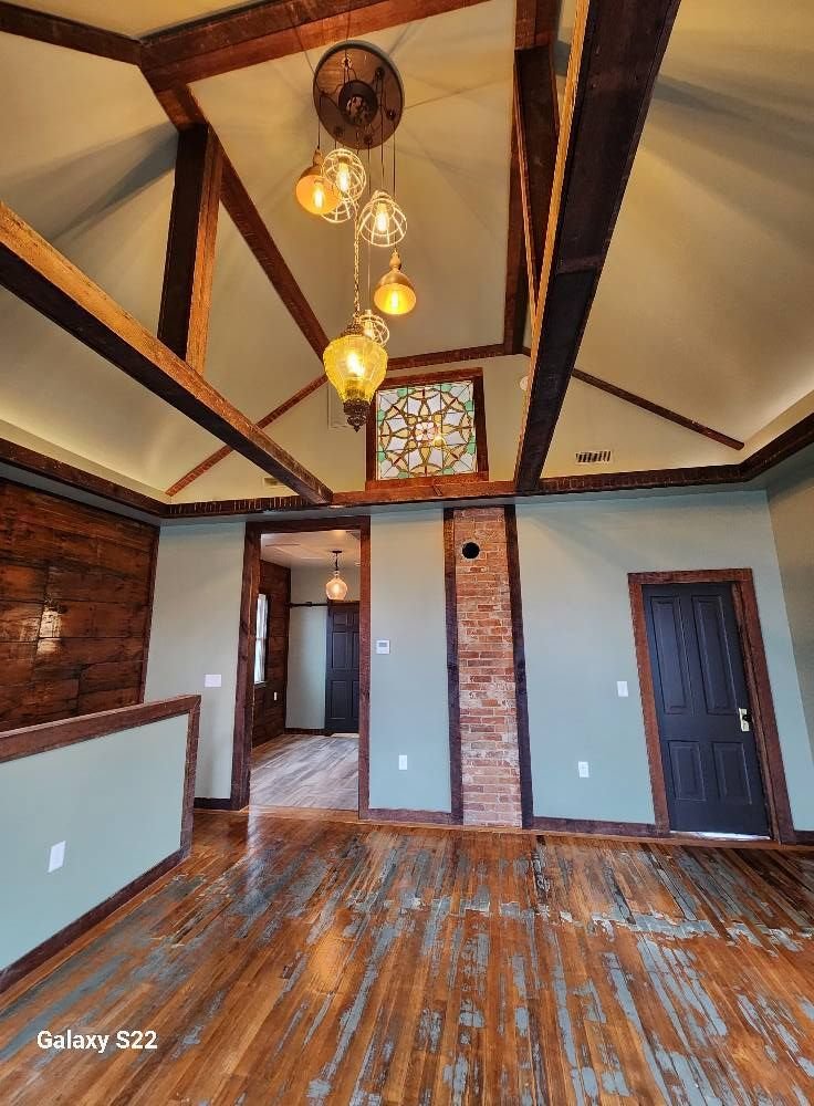 Room with a vaulted ceiling, exposed wood beams, a brick column, hanging pendant lights and worn hardwood floors