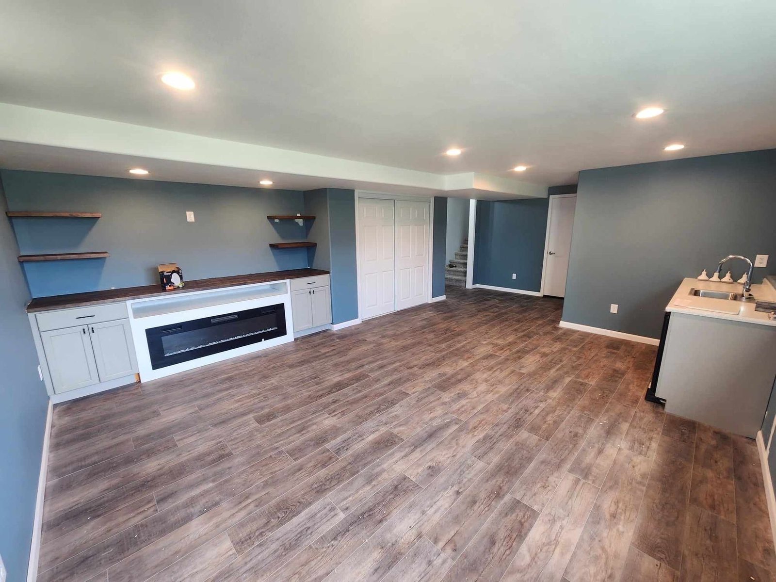 Finished basement room with gray cabinets, built-in electric fireplace, floating shelves, wood-look flooring and recessed lighting