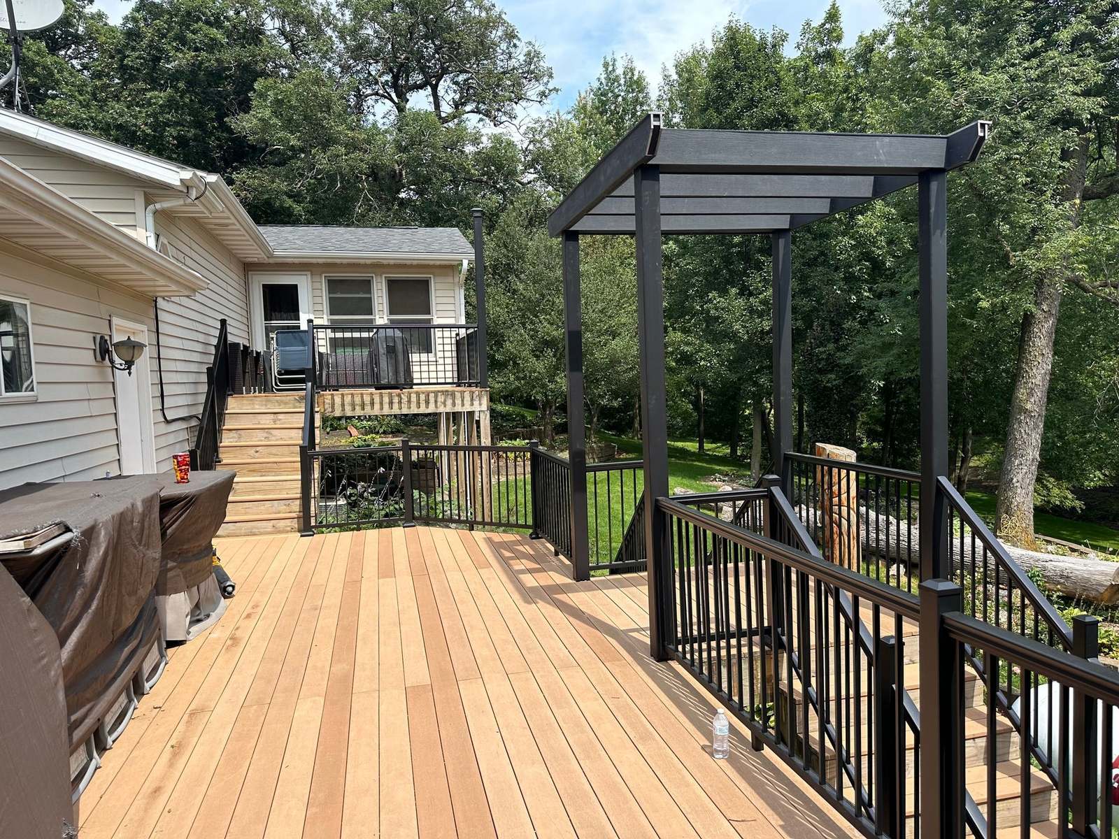 Newly built wood deck with black metal railings and a black pergola beside a house, overlooking trees