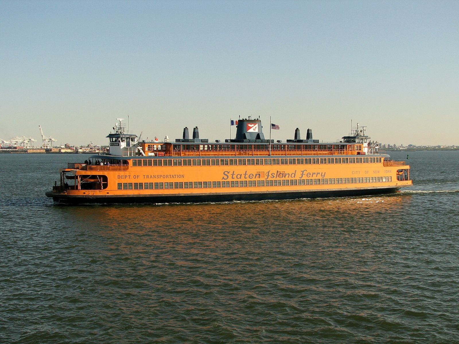 Orange Staten Island Ferry boat crossing calm blue water under a clear sky, with a distant shoreline behind