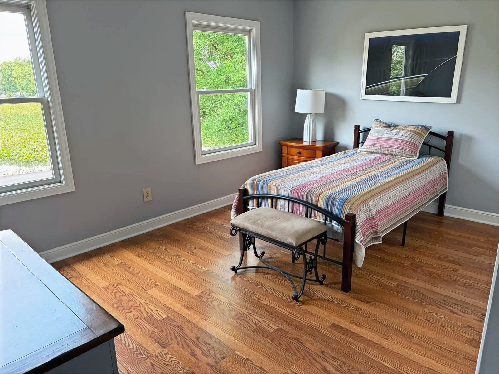 Cleaned bedroom with a striped quilt on a twin bed, polished hardwood floor, and windows looking out on green fields