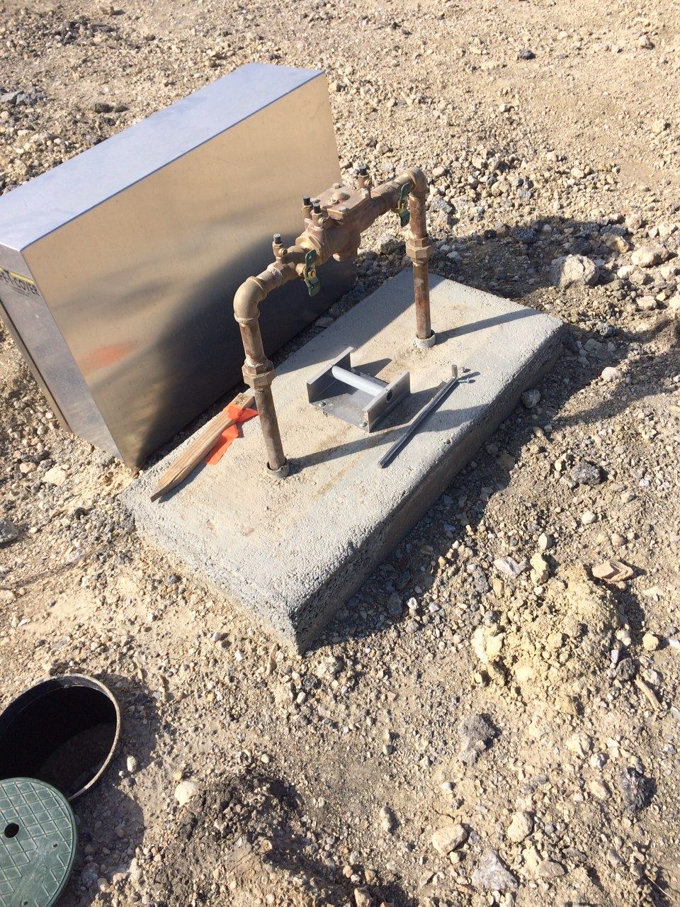 Brass backflow prevention assembly on a concrete pad beside a stainless steel enclosure on a gravel construction site