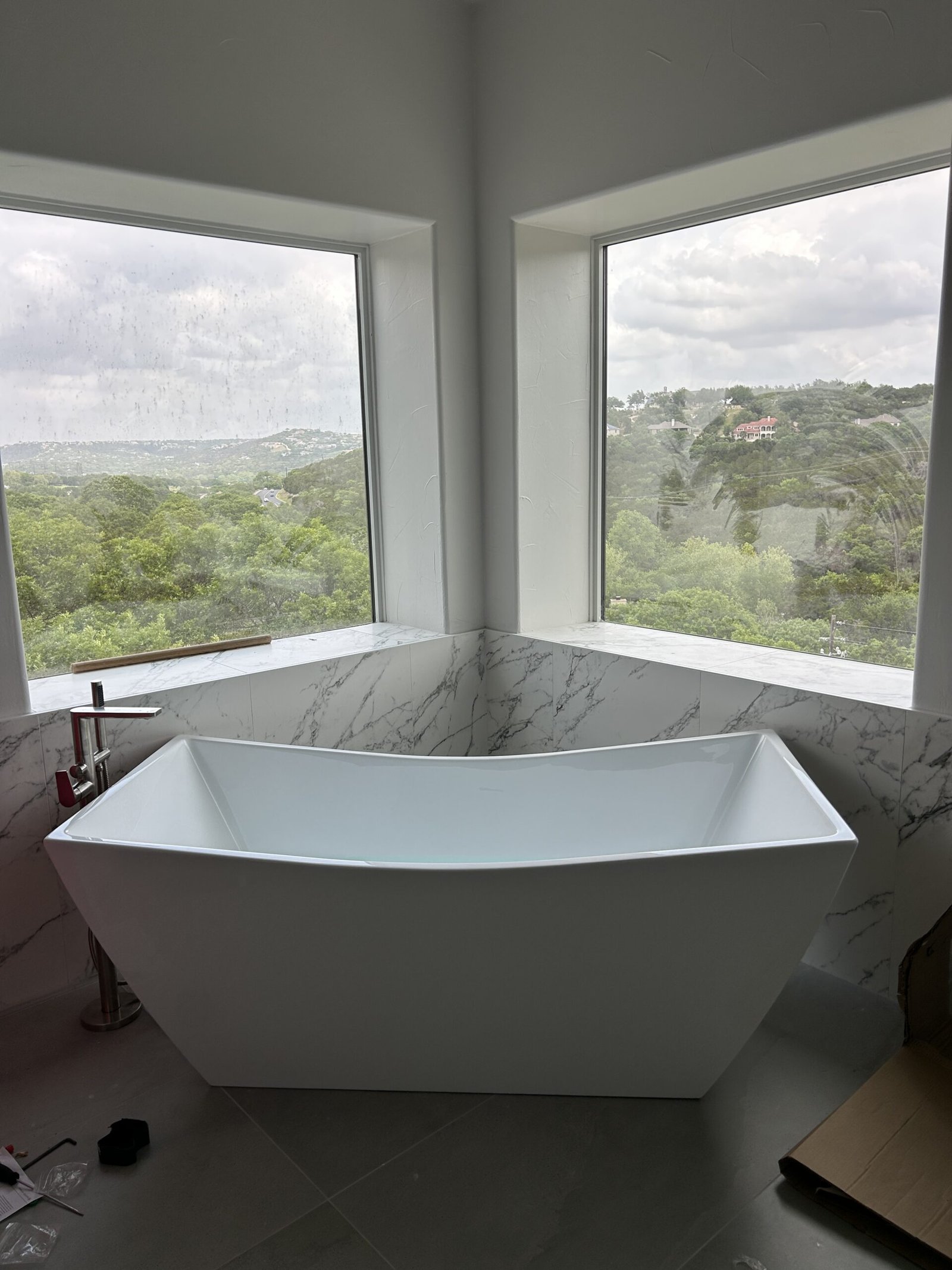 Freestanding white soaking tub in a corner with large windows overlooking green hills and trees