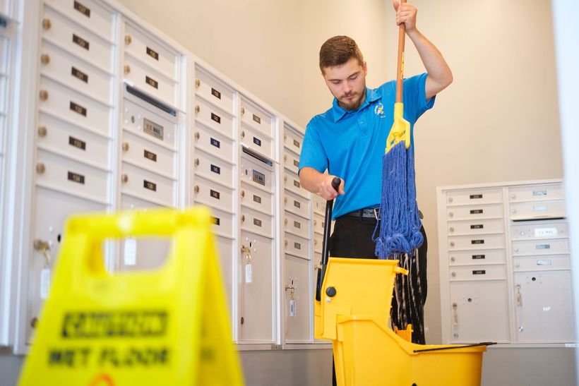 Cleaner in a blue polo wrings a yellow mop over a bucket beside mailboxes and a wet floor sign