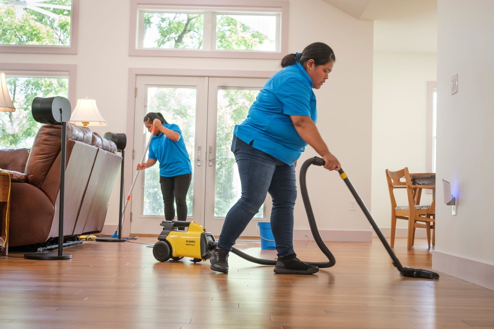 Two cleaners in blue shirts vacuum and dust a bright living room with wood floors