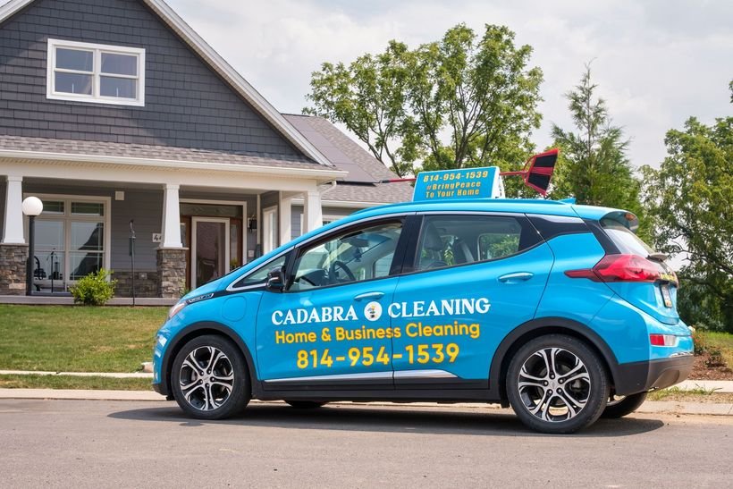 Blue Cadabra Cleaning car with yellow lettering for home and business cleaning, parked on a street in front of a gray house