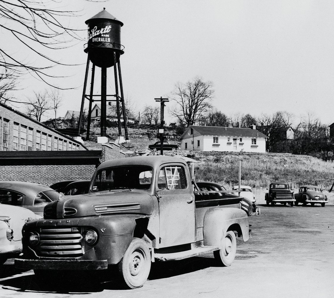 A black-and-white photo of a 1950s pickup truck parked outside a brick factory, with a water tower on the hill behind it painted with the Carhartt name and the word "overalls"