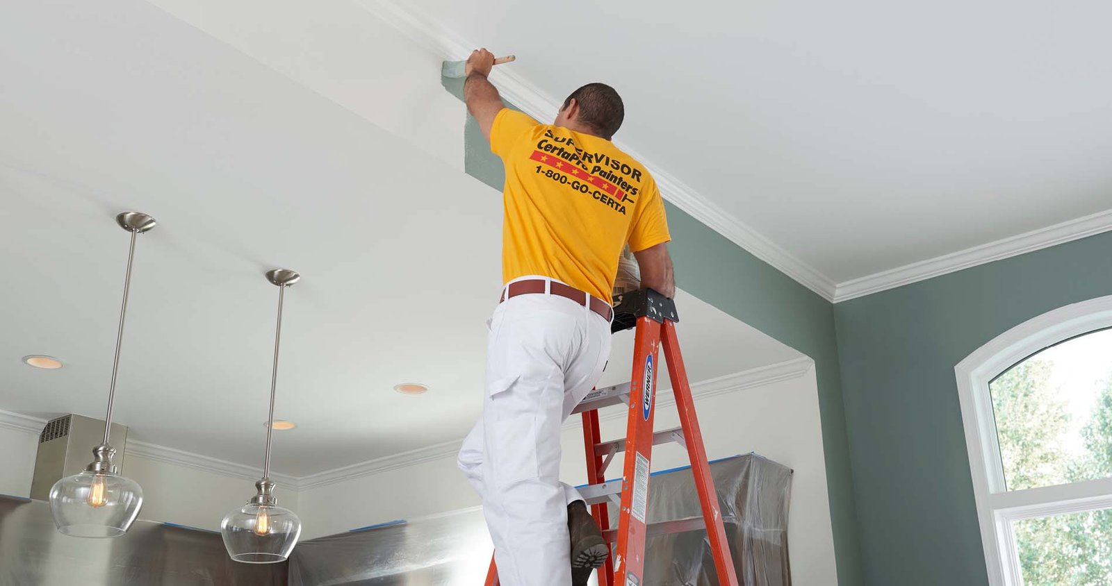 Painter in a yellow CertaPro shirt stands on an orange ladder, brushing paint along the ceiling edge of a room