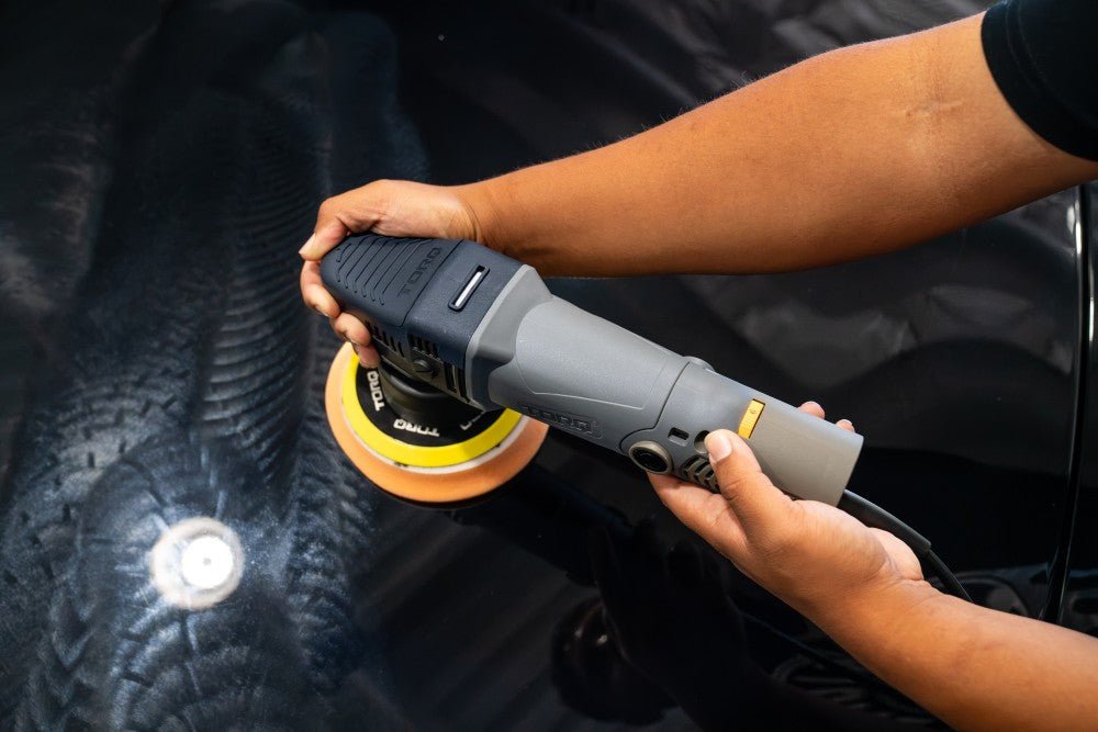 Hands guiding a dual-action polisher with an orange foam pad across a dark car panel