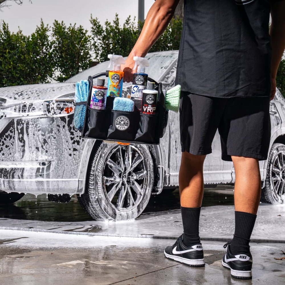 Person holding a black caddy of Chemical Guys bottles and towels beside a car covered in white foam