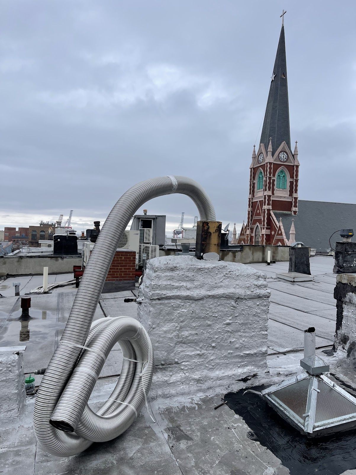 A coil of flexible stainless steel chimney liner beside a chimney on a city rooftop, with a church steeple in the background