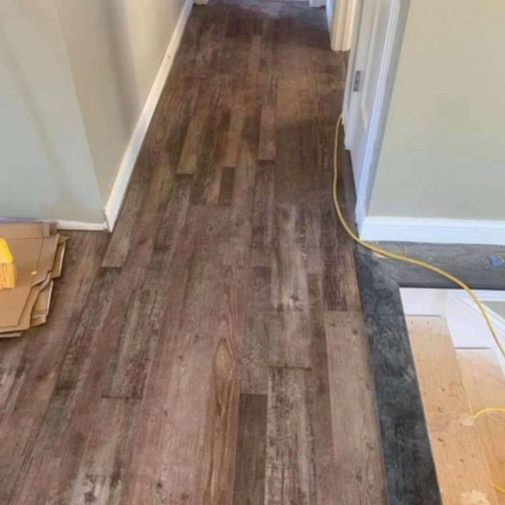 Wood-look plank flooring installed down a hallway, with a yellow cord running along one wall and bare plywood at the edge