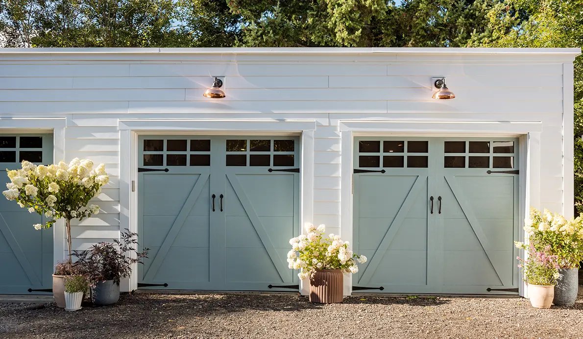 Two pale blue carriage-style garage doors with X-brace panels and black hardware on a white building, flanked by flowering planters