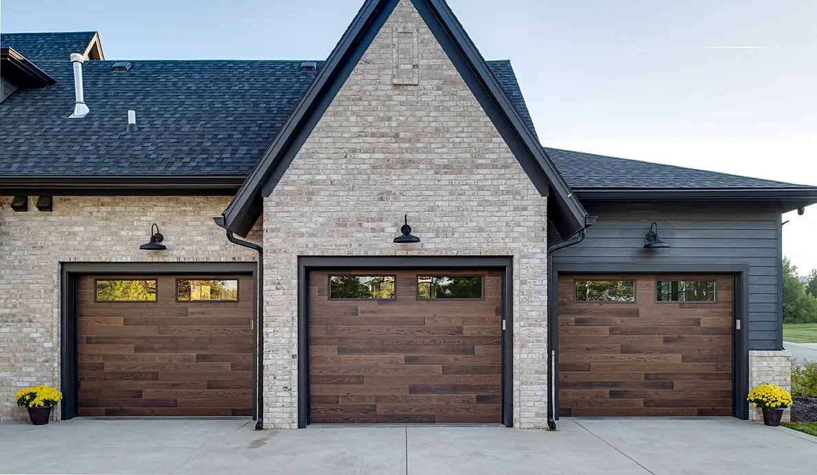 Three brown wood-look garage doors with small top windows set in a light brick gable with black trim and gooseneck lights