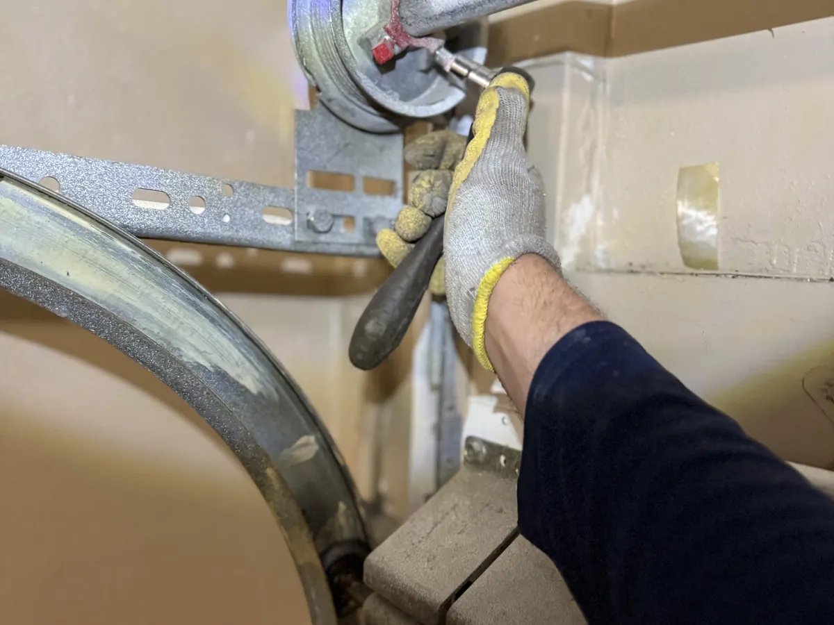 Technician in a gloved hand uses a tool on a cable drum and bracket of a garage door