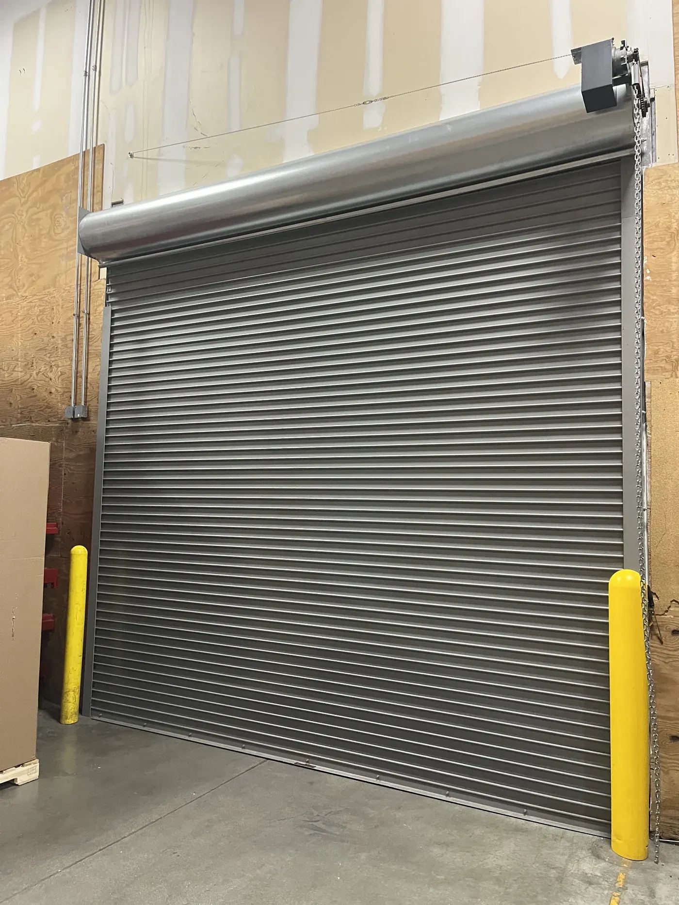 Gray steel rolling door with chain hoist and yellow safety bollards inside a warehouse loading area