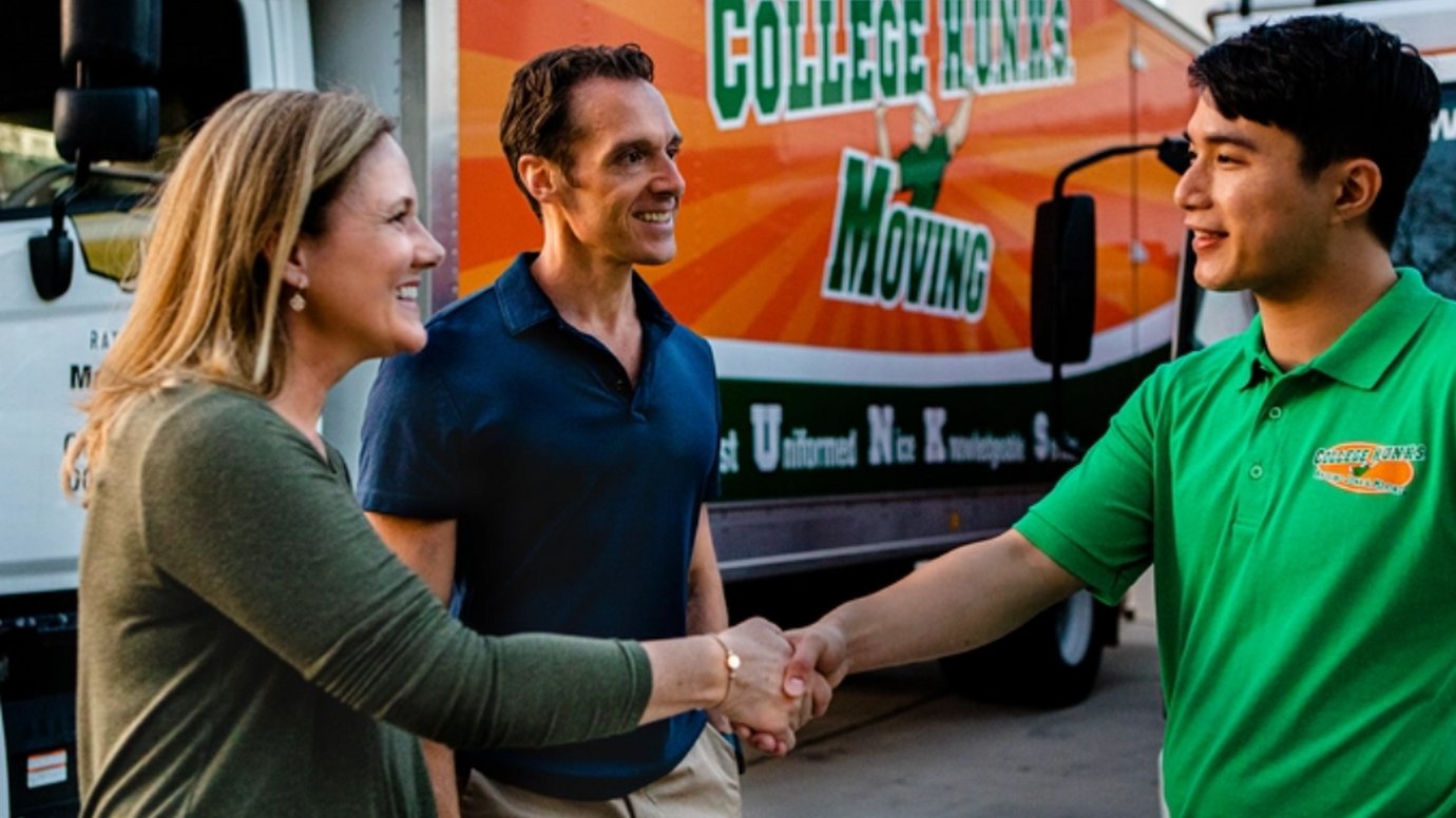A College Hunks mover in a green polo shakes hands with a smiling woman beside a branded moving truck
