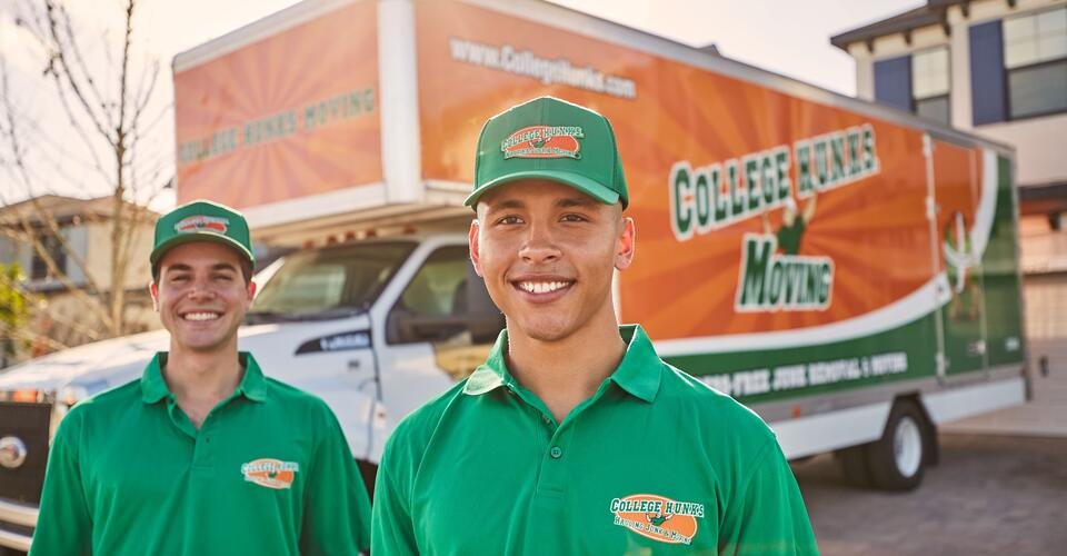 Two smiling crew members in green College Hunks polos and caps stand in front of an orange and green moving truck