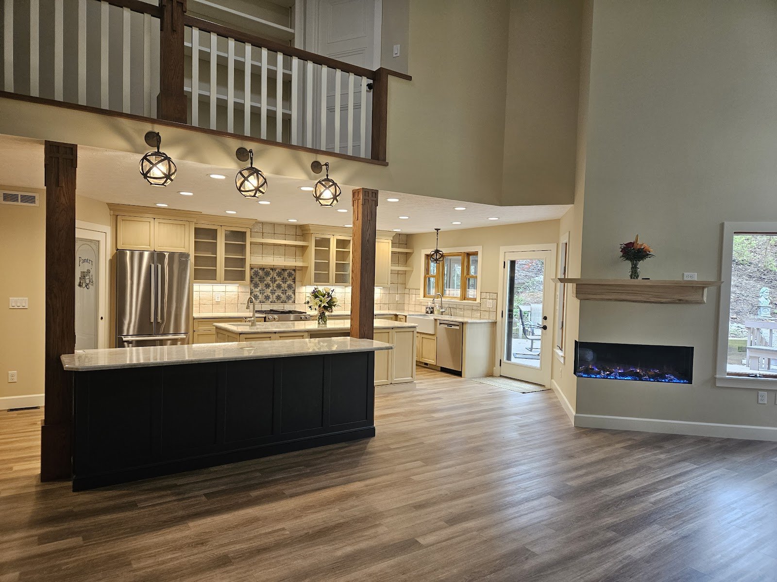 Open-plan remodeled kitchen with a dark island, glass-front cabinets, exposed beam posts and a linear fireplace under a loft