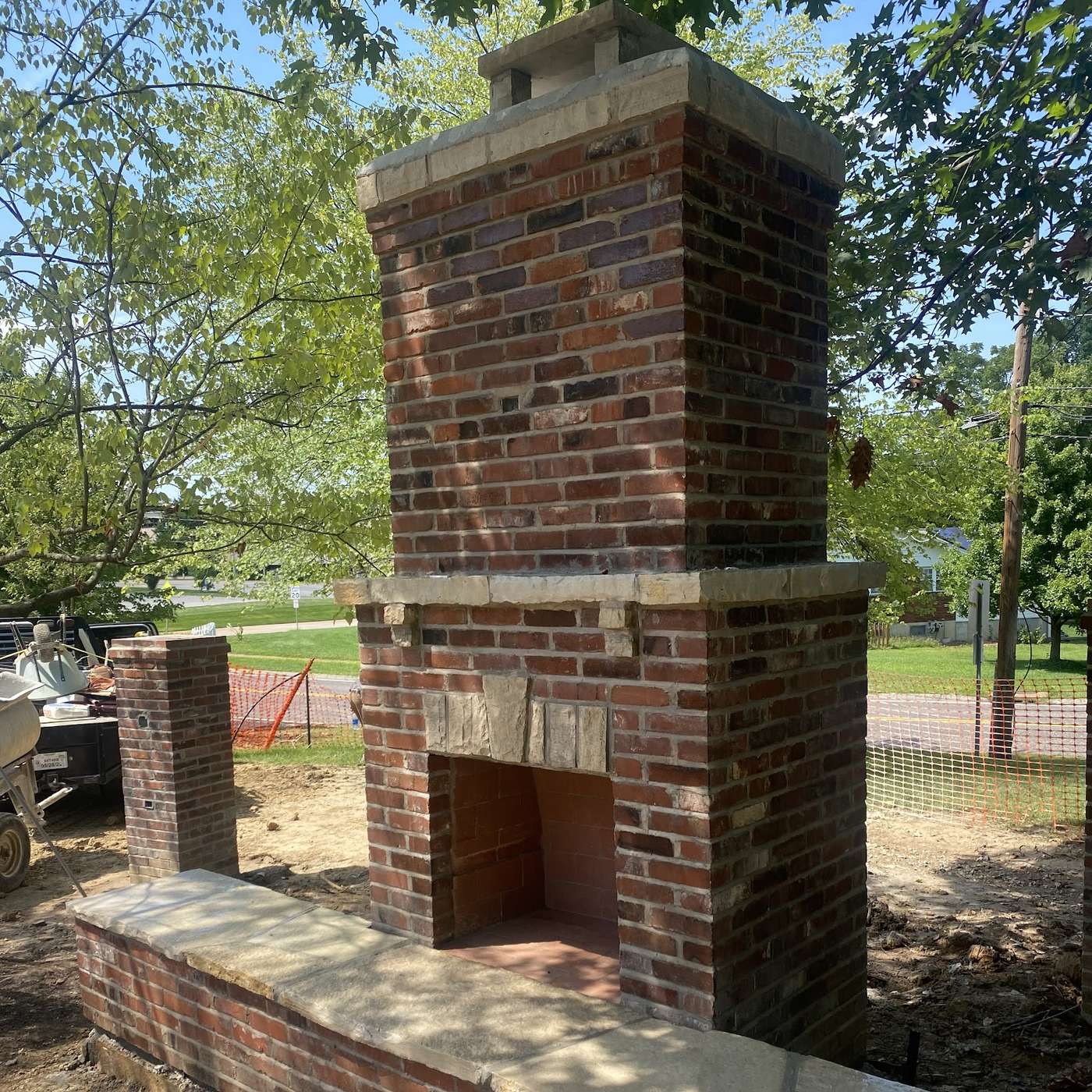 A brick outdoor fireplace with a tall chimney rising from a low brick bench wall, on a bare dirt site with a truck parked behind it