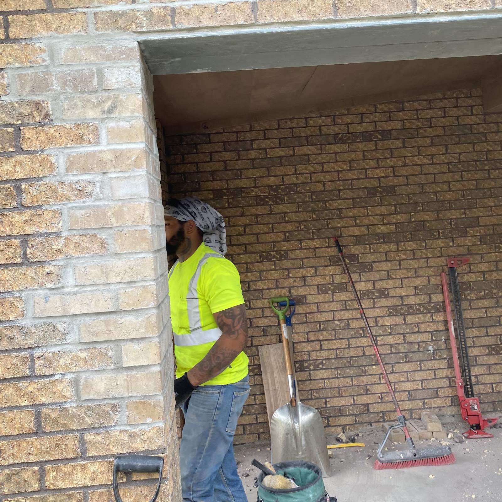 A worker in a yellow safety shirt and head wrap stands inside a brick opening, with a shovel, a broom and a jack behind him