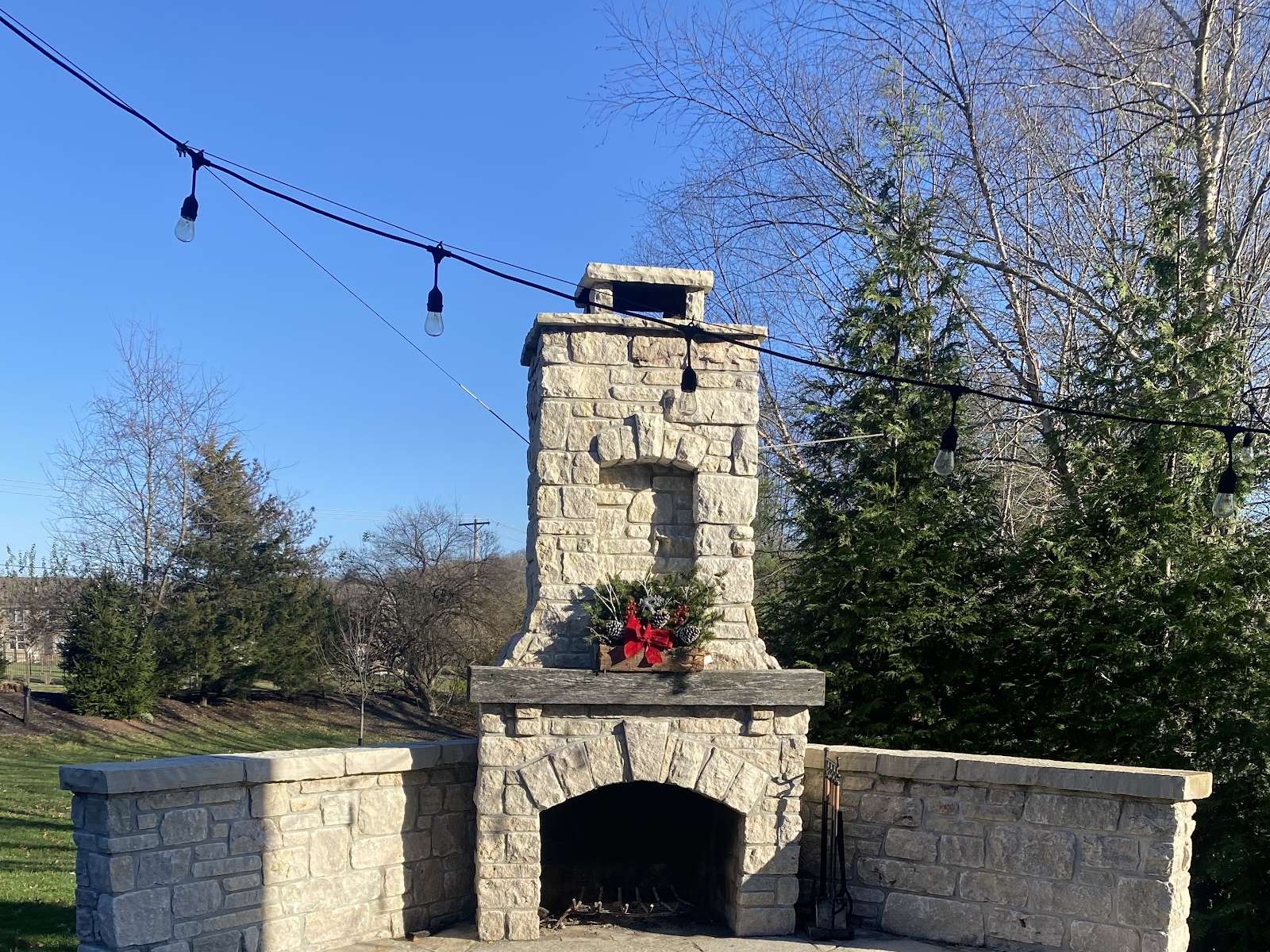 A stone outdoor fireplace with a tall chimney and low stone seating walls, under a string of patio lights